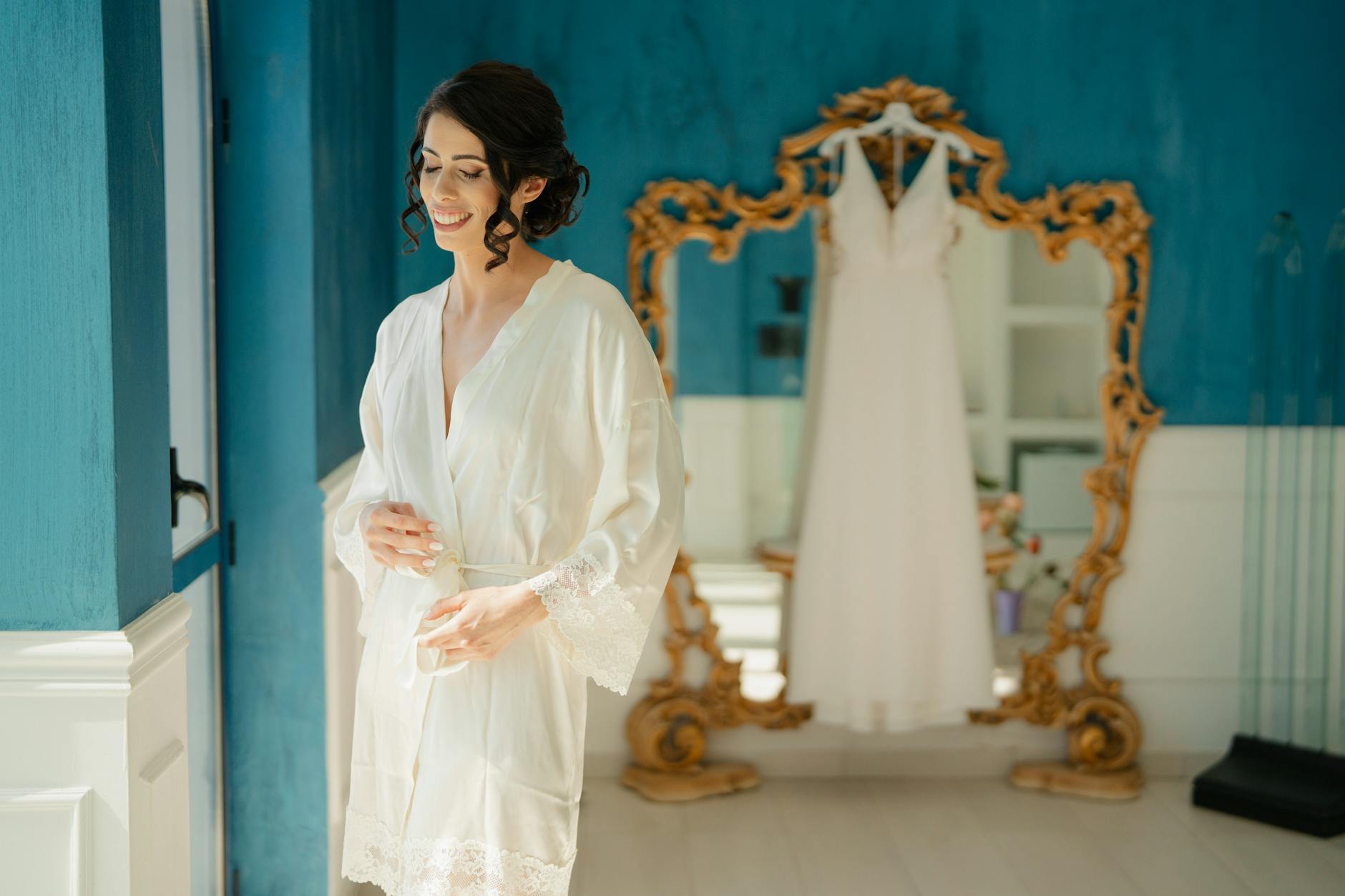 A bride in a robe smiles while preparing for her wedding in a stylish blue room with a gold-framed mirror.
