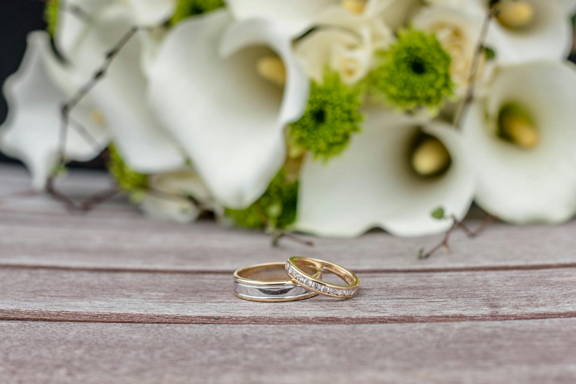 Golden wedding rings placed on wooden surface near fragrant bouquet of delicate white arum lilies