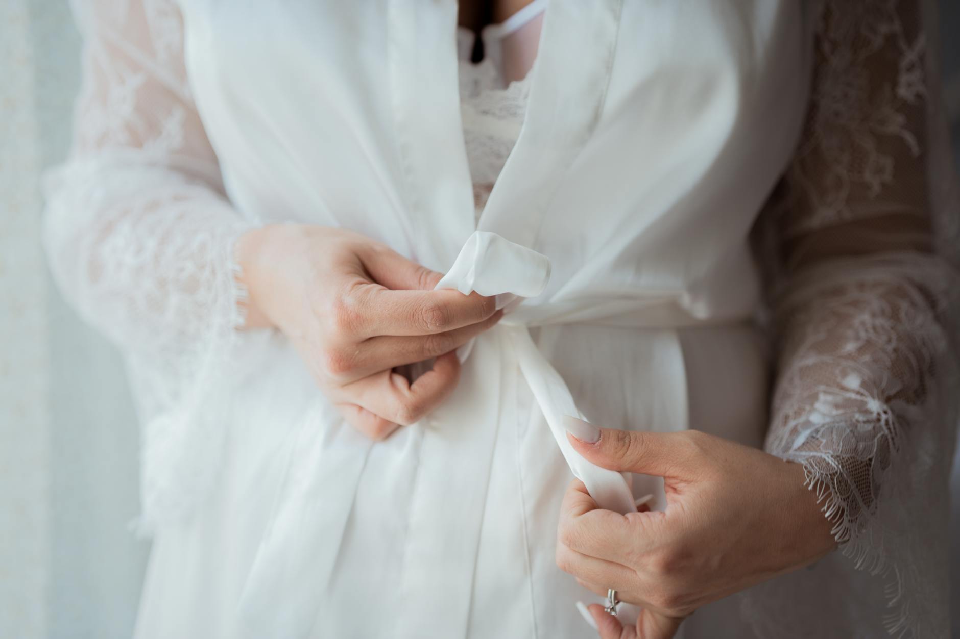 Close-up of a woman delicately tying the belt on an elegant white lace robe. Soft and intimate detail shot.