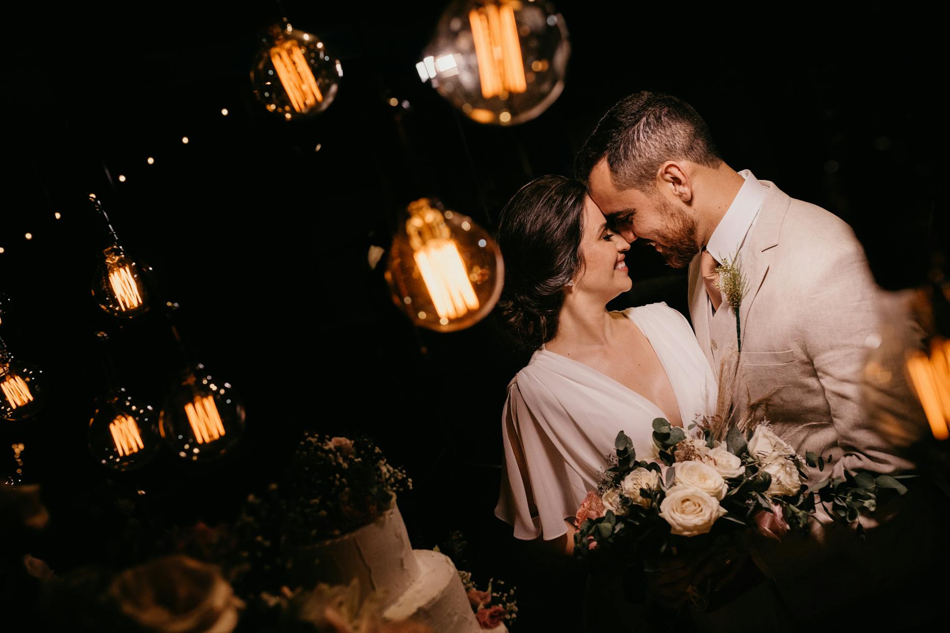Bride and groom sharing a tender moment under warm pendant lights with a bouquet of flowers.