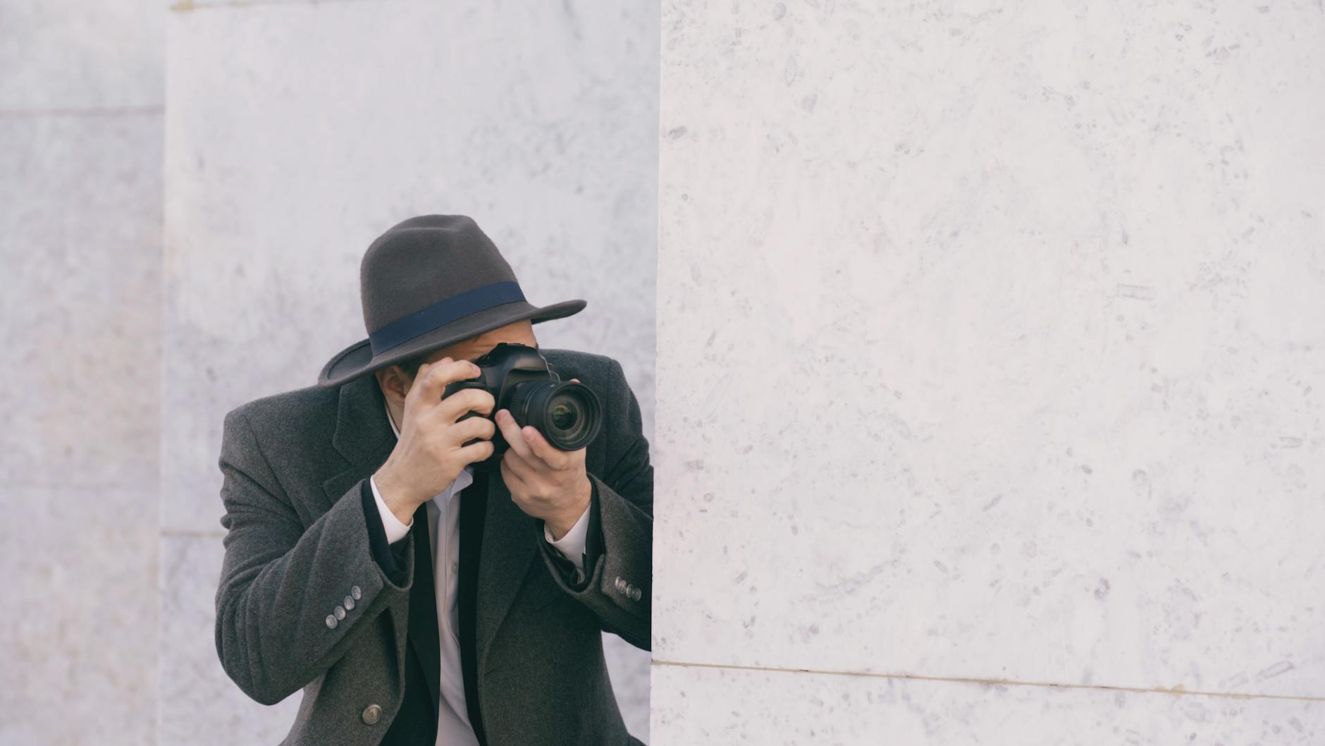 A person with a camera and gray hat capturing photos against a marble wall.
