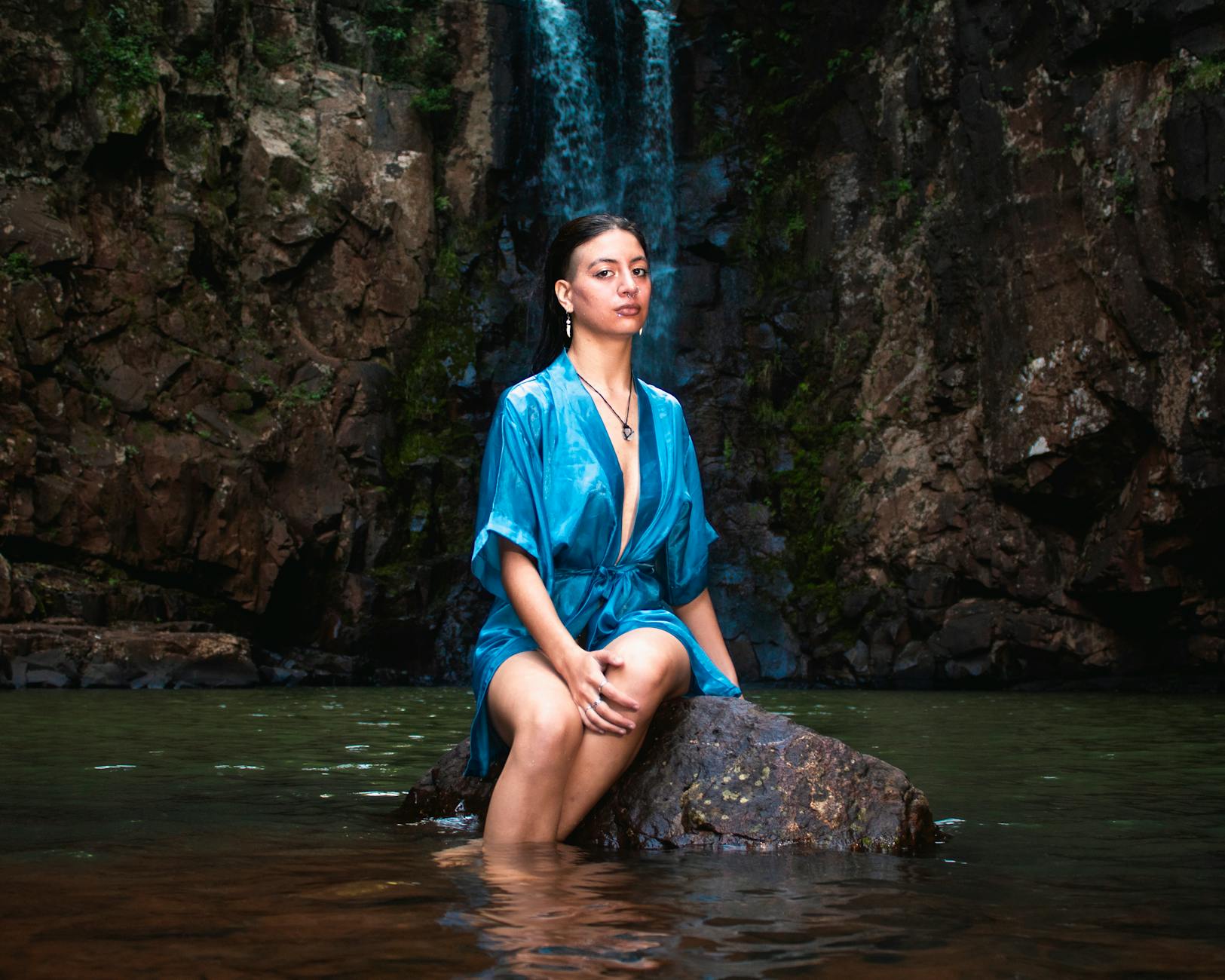 Portrait of a woman in a blue gown posing by a scenic waterfall.
