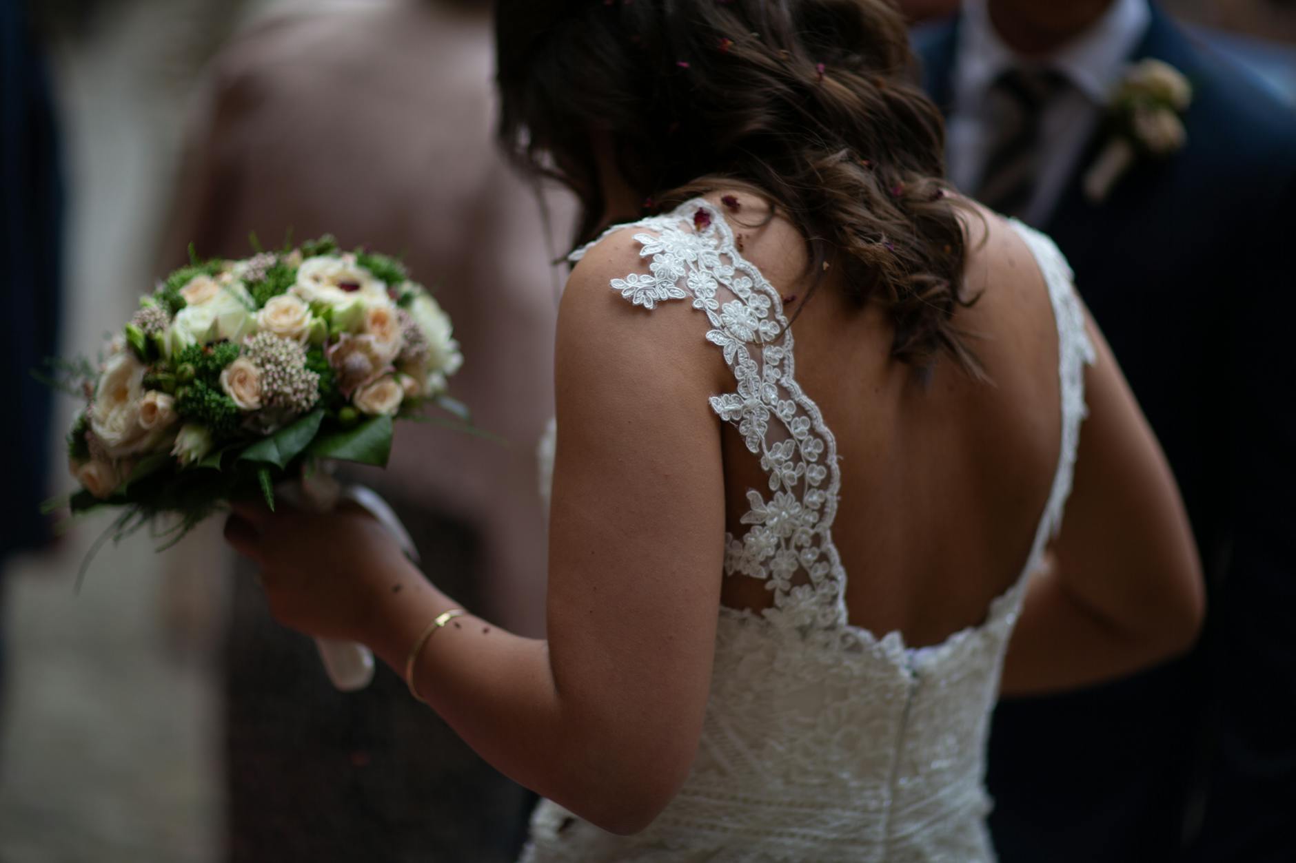 A bride in a lace gown holds a beautiful bouquet, viewed from behind at a wedding ceremony.