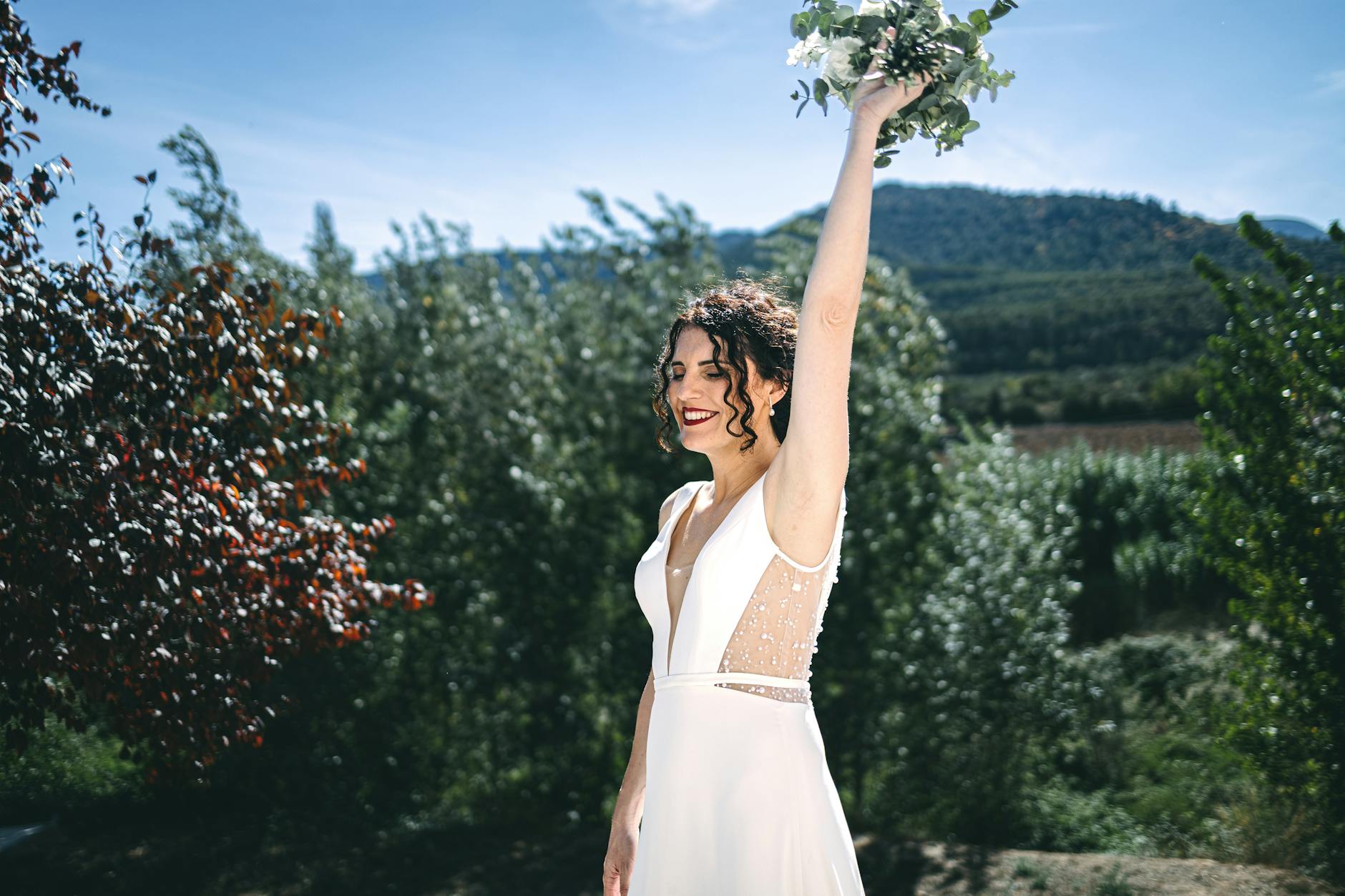 A smiling bride raises her bouquet in a vibrant outdoor setting, embracing nature and joy.
