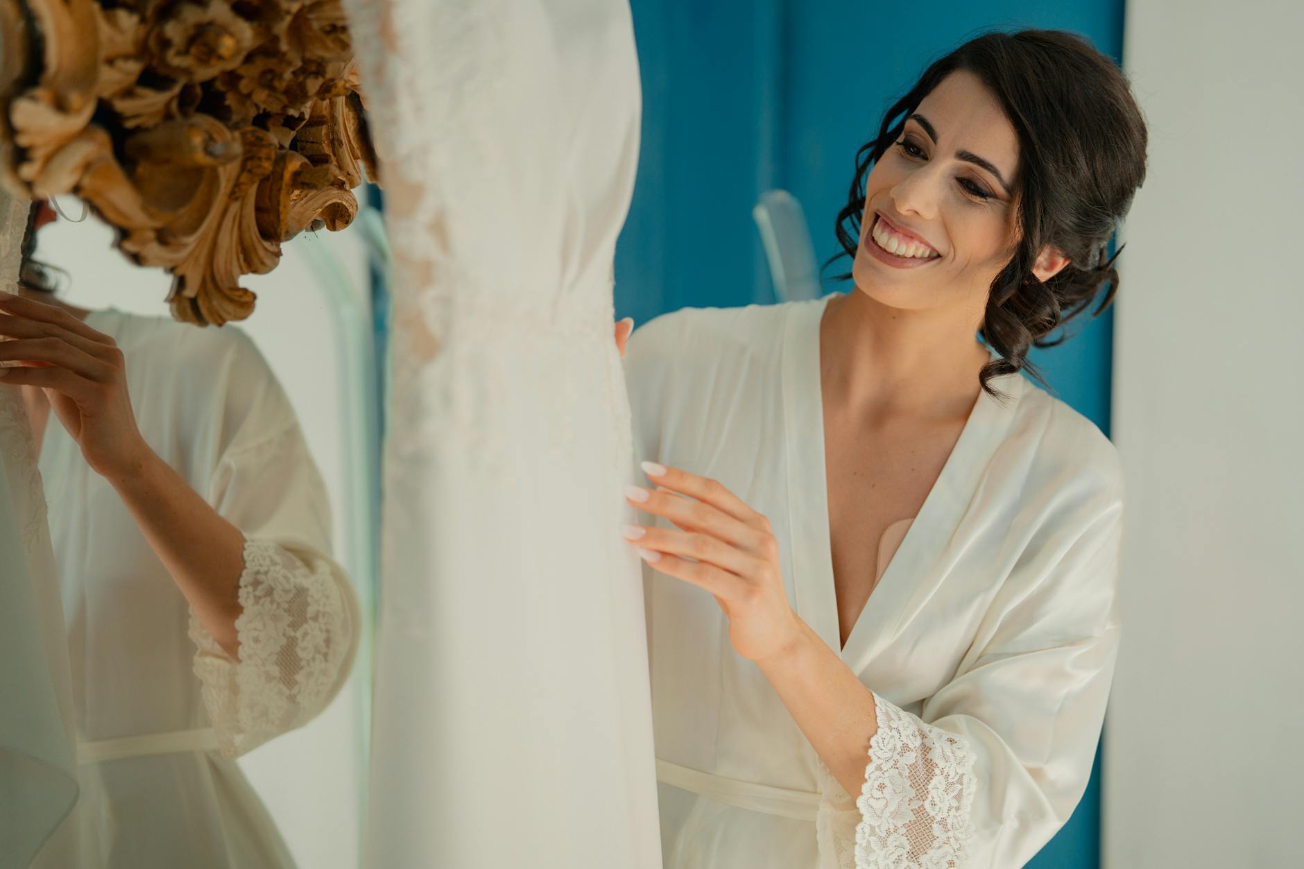 Bride in robe smiling while looking at a wedding dress, preparing for the big day indoors.