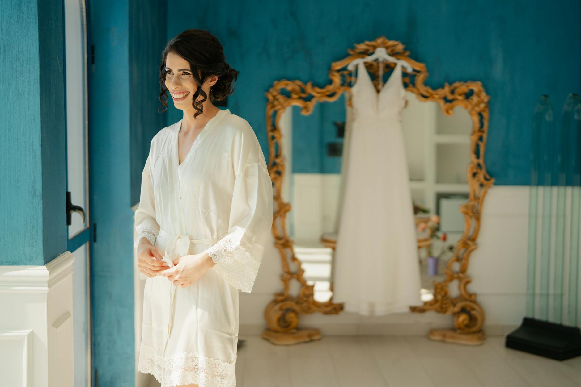 A bride in a white robe stands smiling in an elegant room with her wedding dress hanging on a mirror.